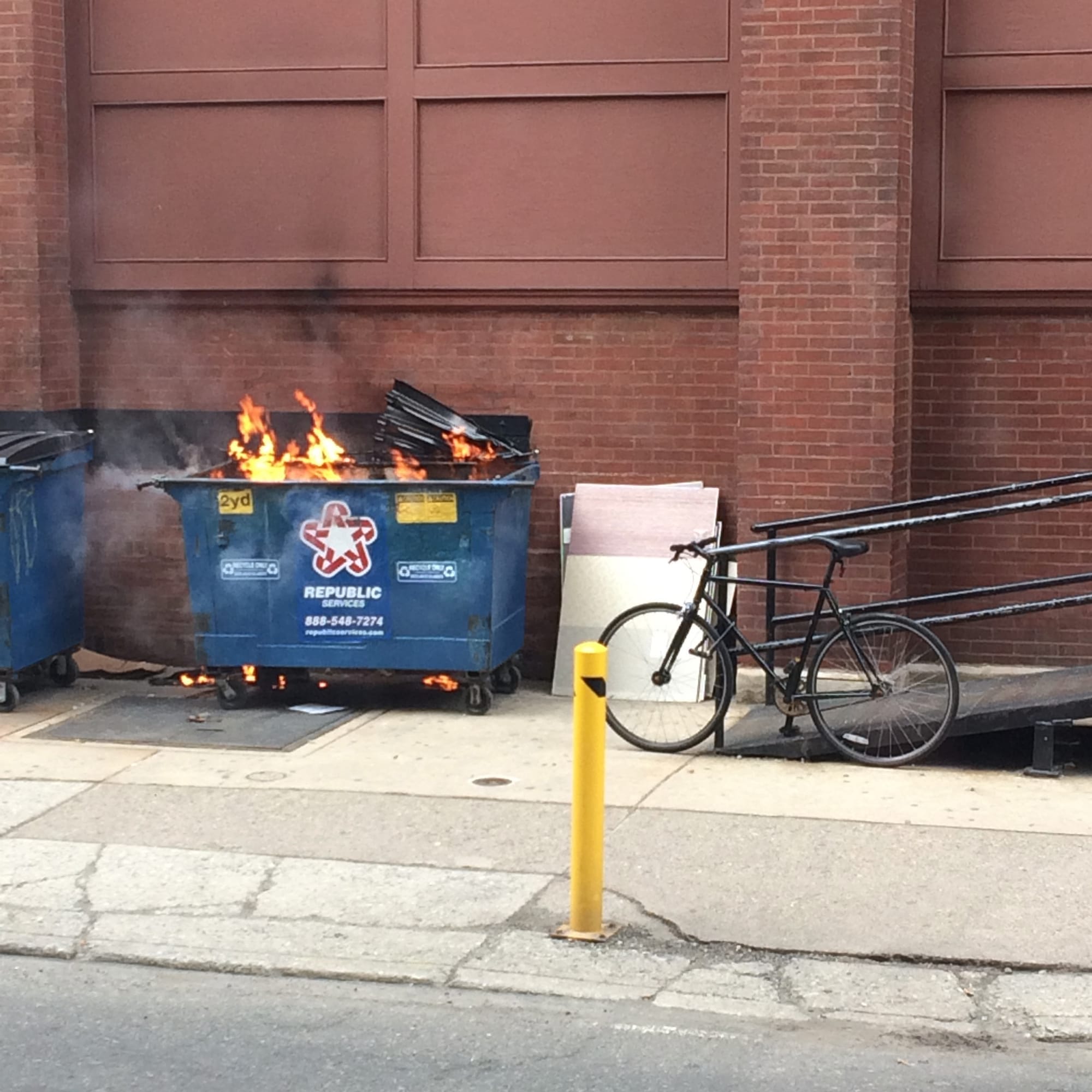 A city street showing a blue trash dumpster with bright flames rising out of it. A black bicycle is parked next to it.