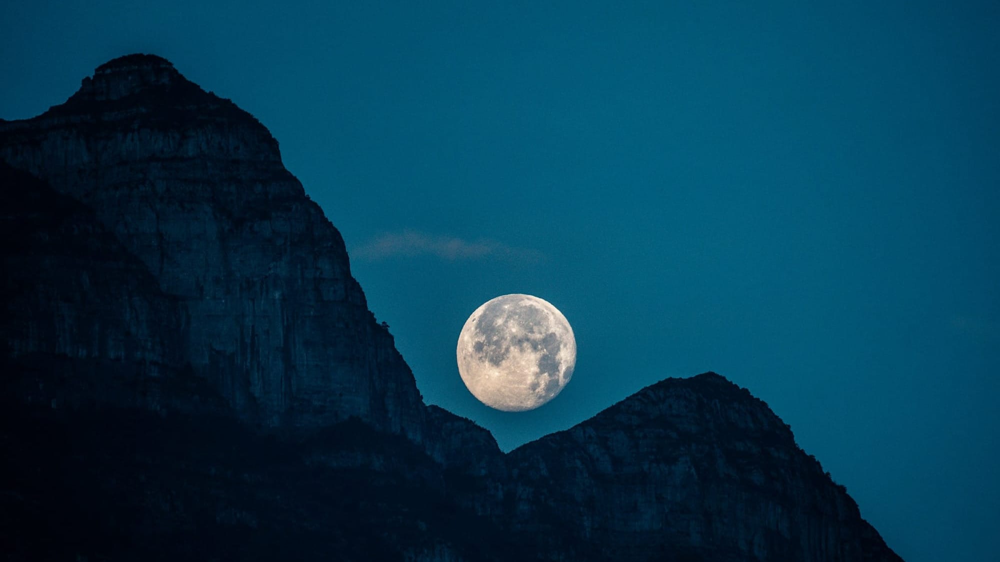 A full moon hangs in a turquoise sky above the outline of a craggy mountain range.